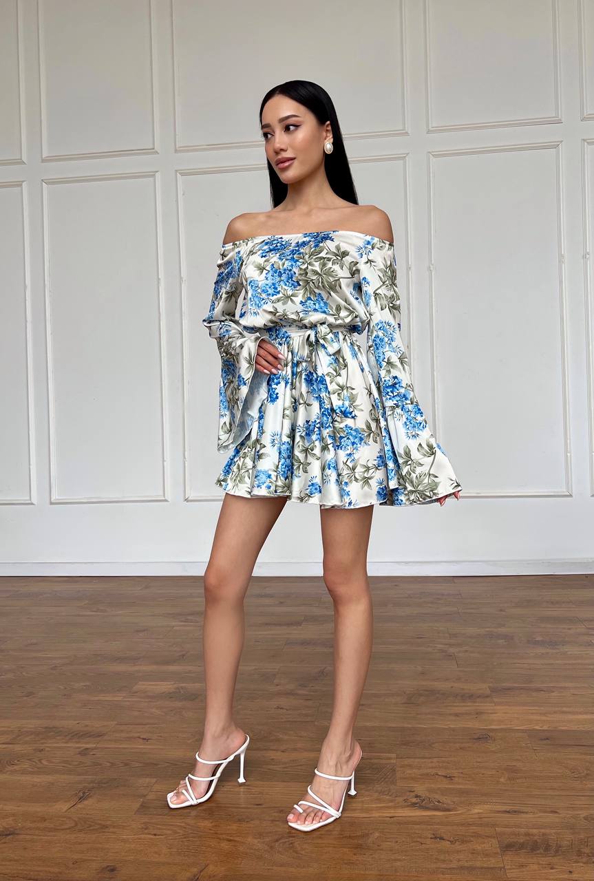 Woman wearing a floral off-shoulder dress standing against a white paneled wall.