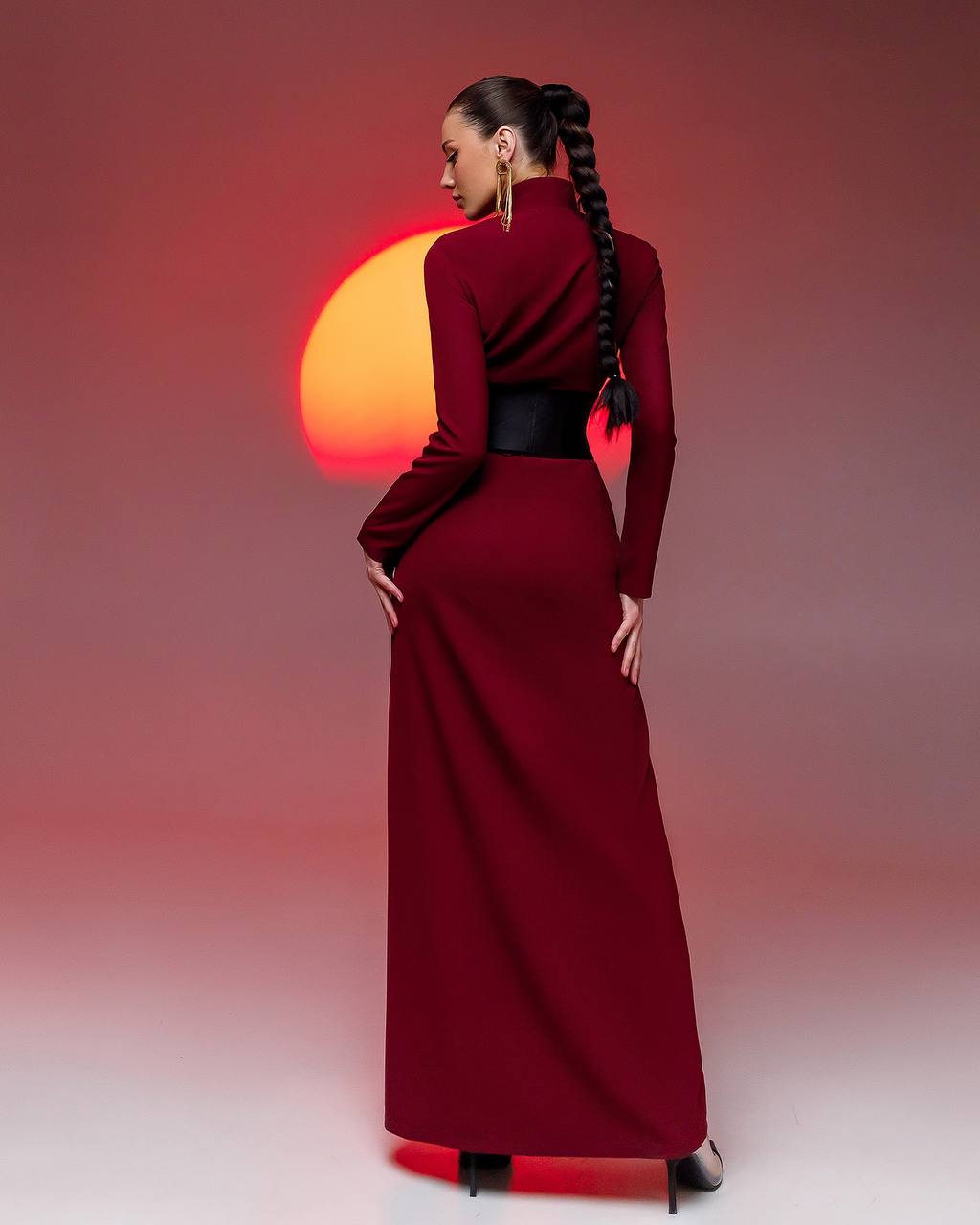 Woman in a long red dress with a black belt against a gradient background with a sun-like shape.