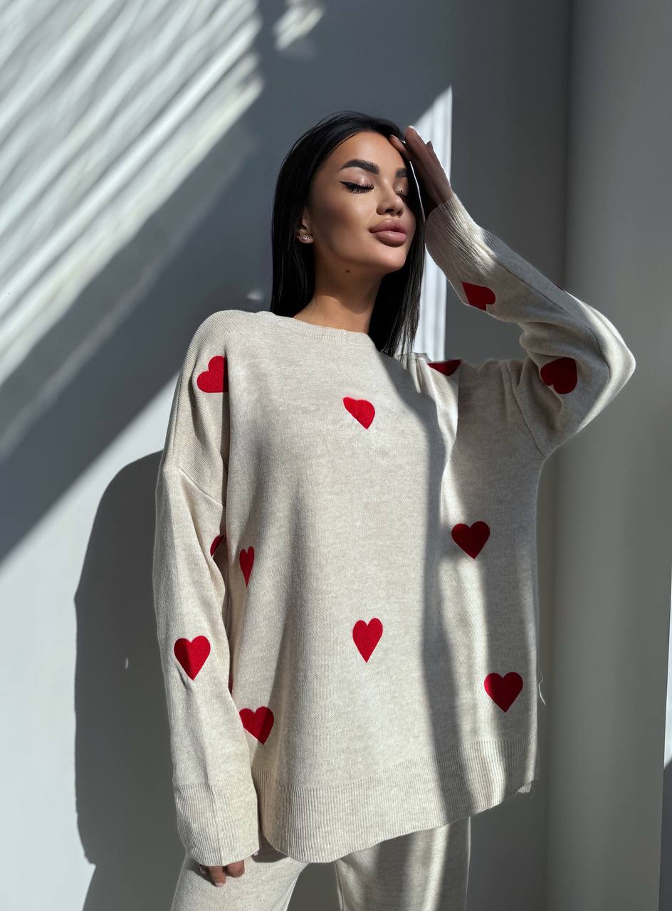 Woman wearing a cream sweater with red heart patterns against a white wall.
