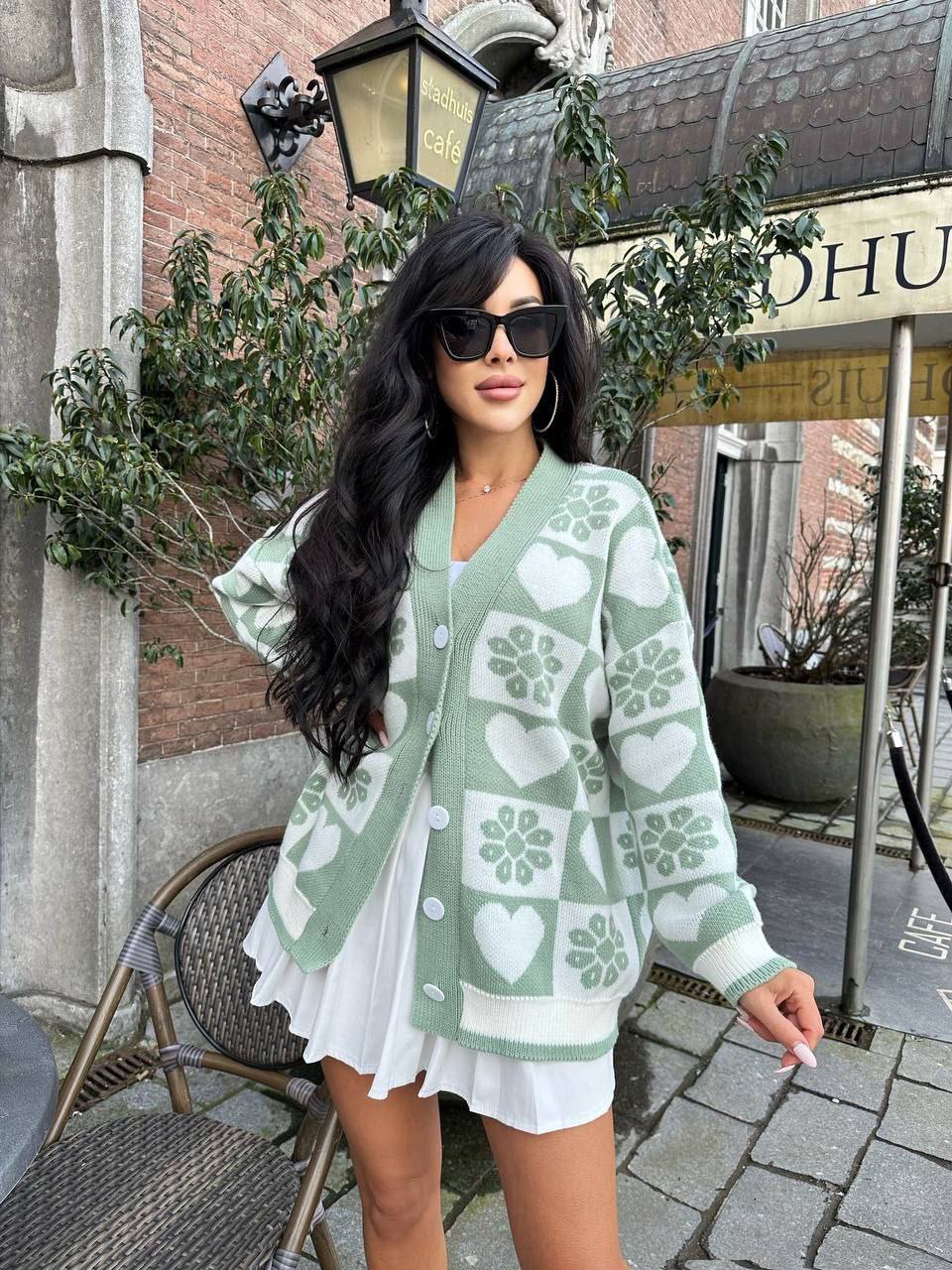 Woman wearing a green patterned cardigan outdoors with a building and sign in the background.