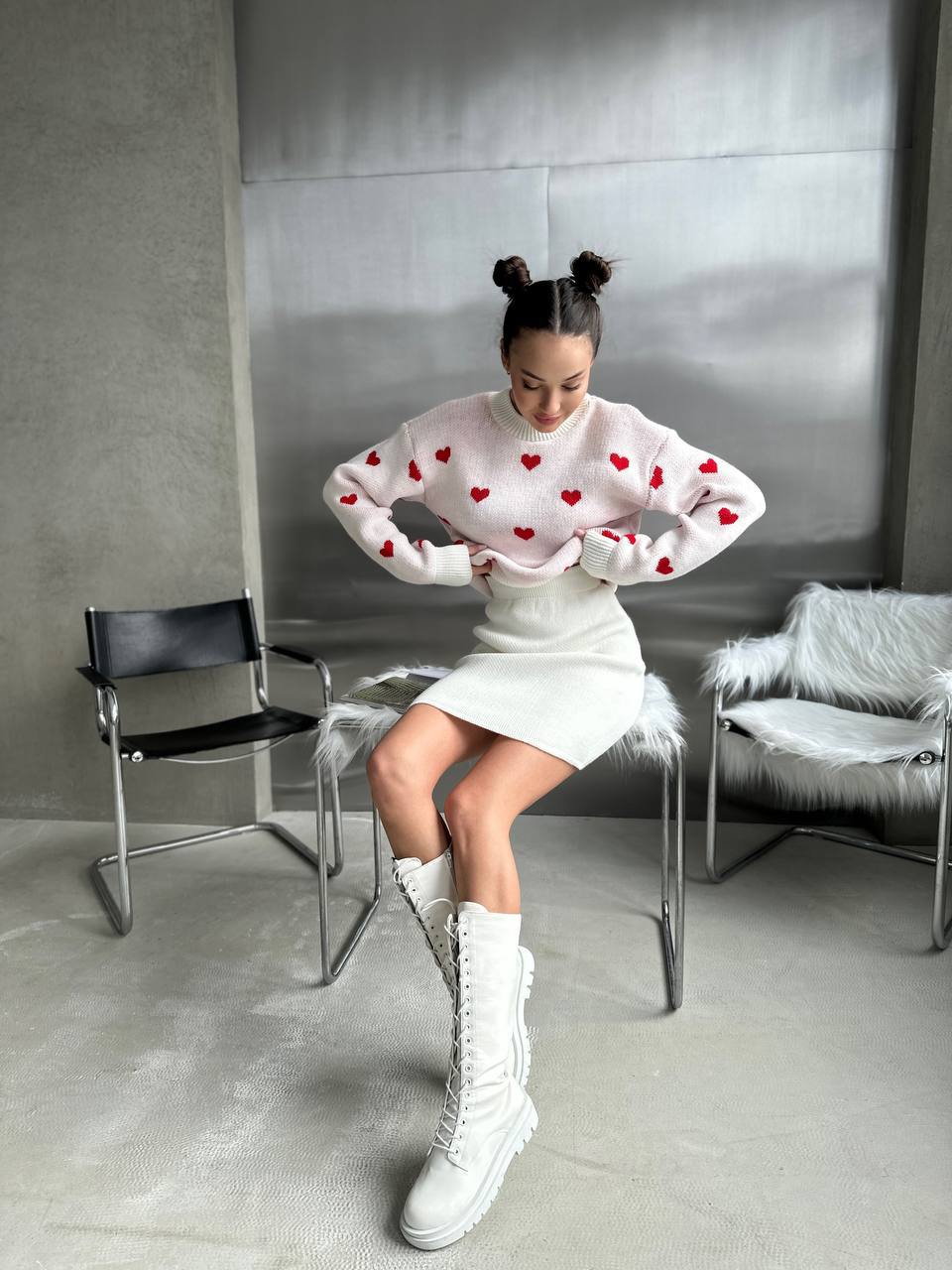 Woman wearing a white sweater with red heart patterns, white skirt, and white boots in a modern room.