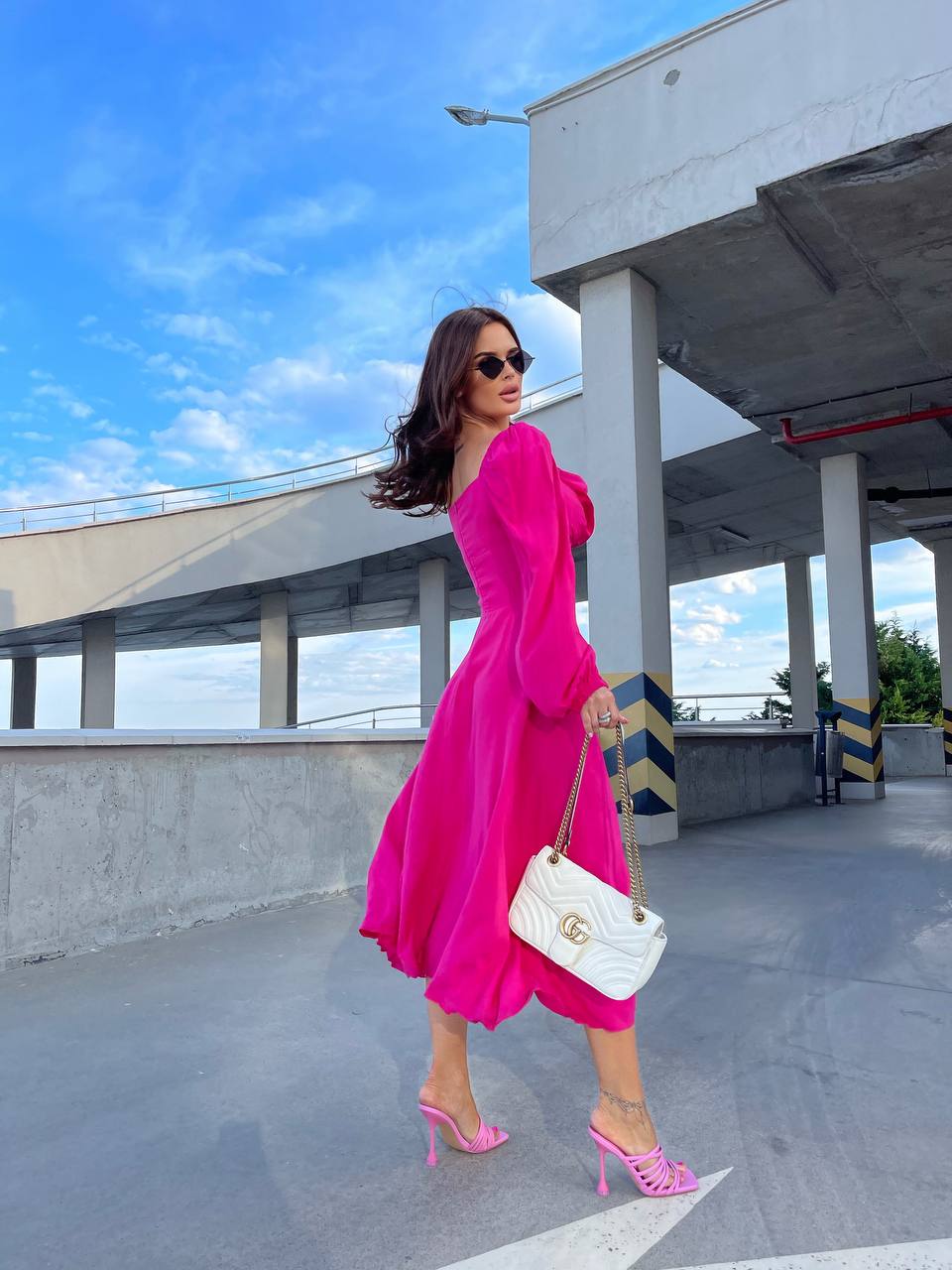 Woman in a bright pink dress with matching shoes holding a white handbag on a rooftop.