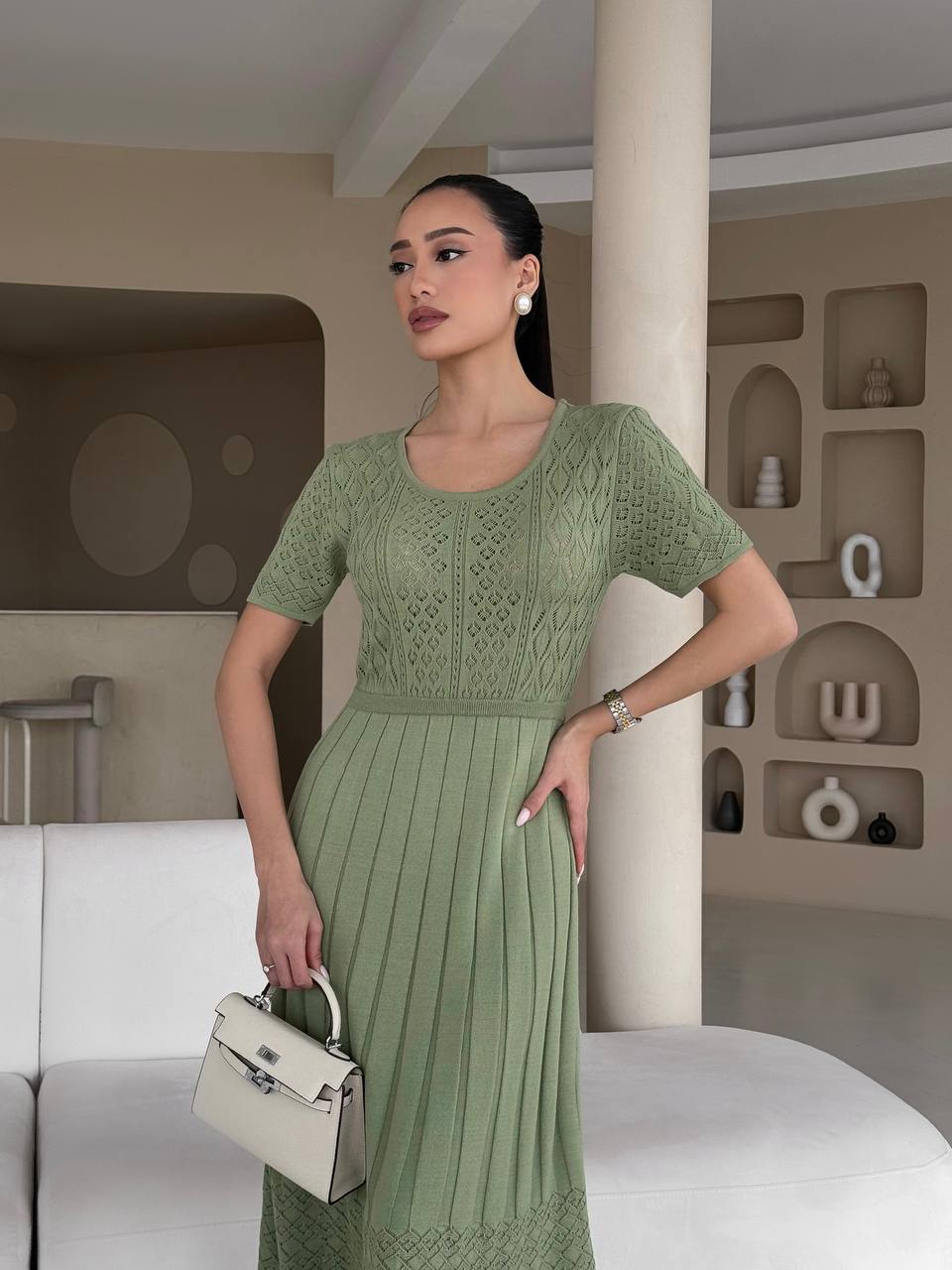 Woman in a green dress holding a white handbag in an indoor setting.