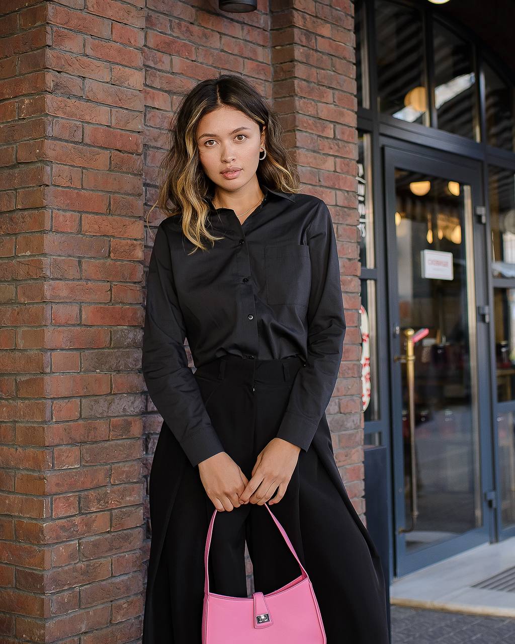 Woman in black outfit holding a pink handbag against a brick wall.