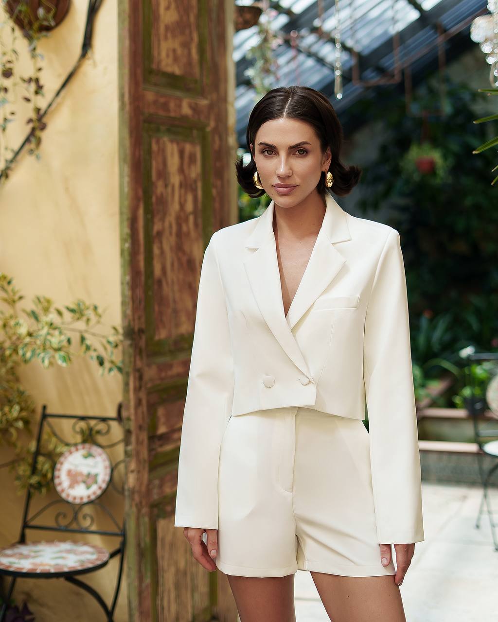 Woman in a white suit standing in an outdoor setting with plants and a decorative wall.