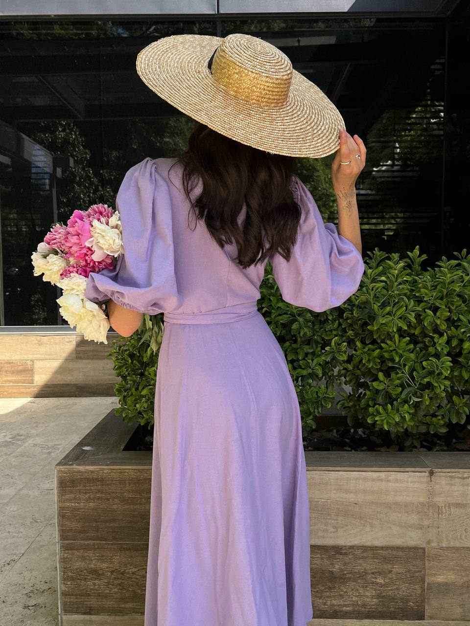 Woman in a lavender dress and straw hat holding flowers, standing outdoors.