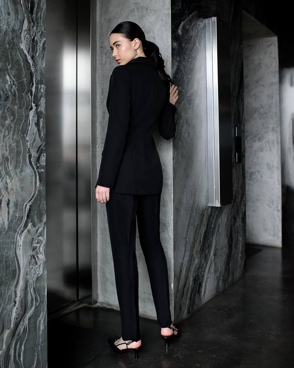 Woman in a black suit standing in a modern interior setting with marble walls.