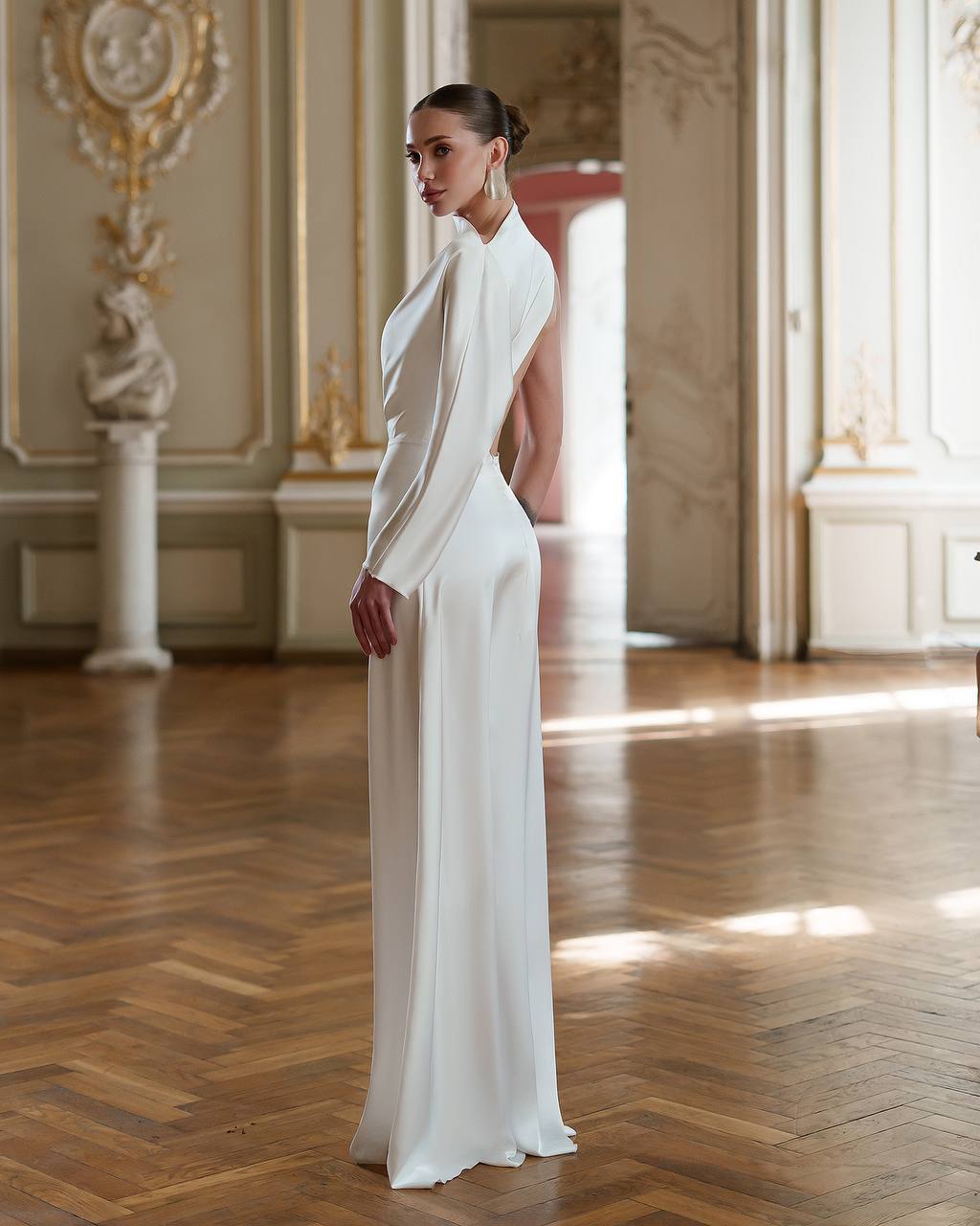 Woman in a white evening gown standing in an elegant room with wooden floors and decorative walls.