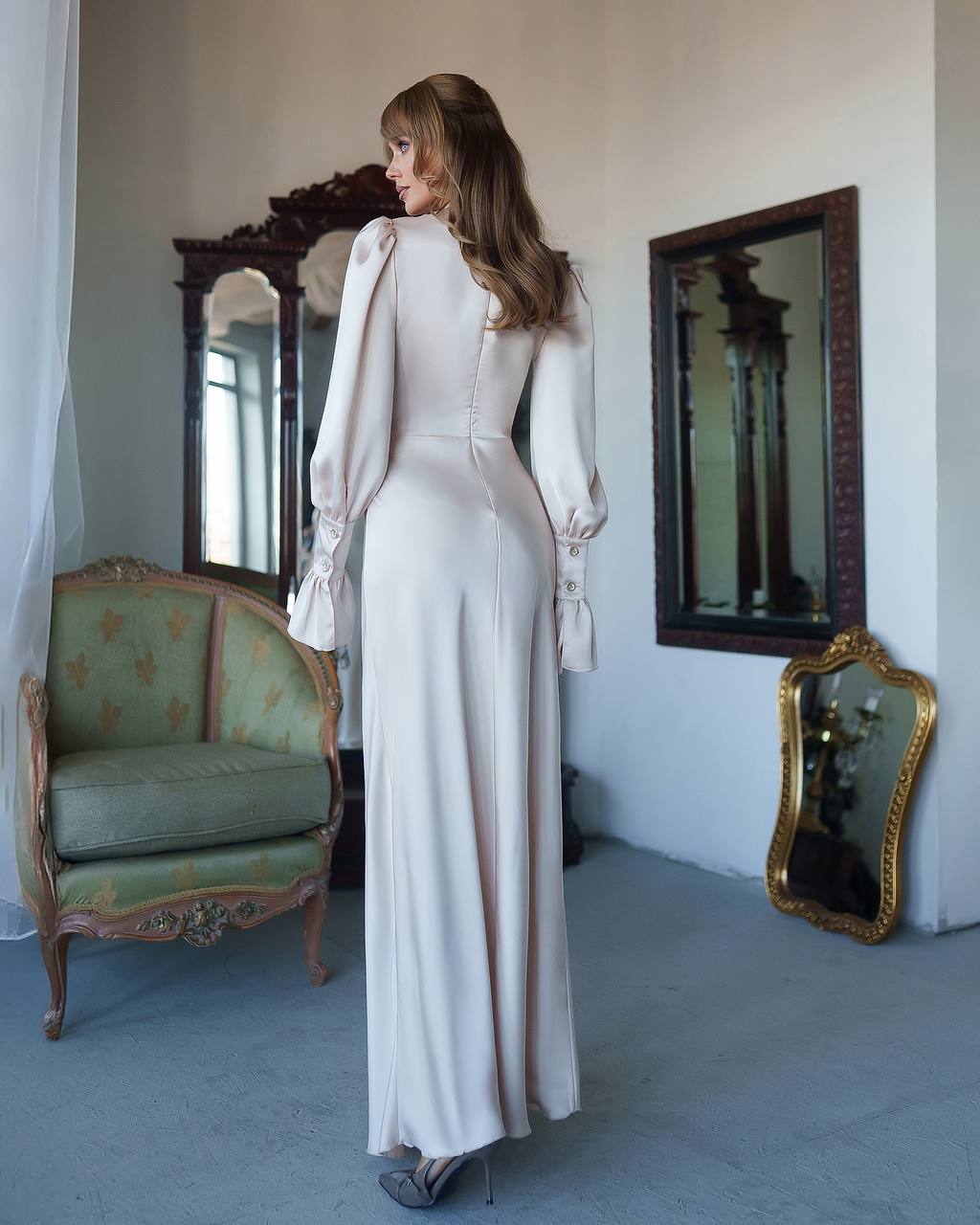 Woman in a long, light-colored dress standing in an elegant room with mirrors and a chair.