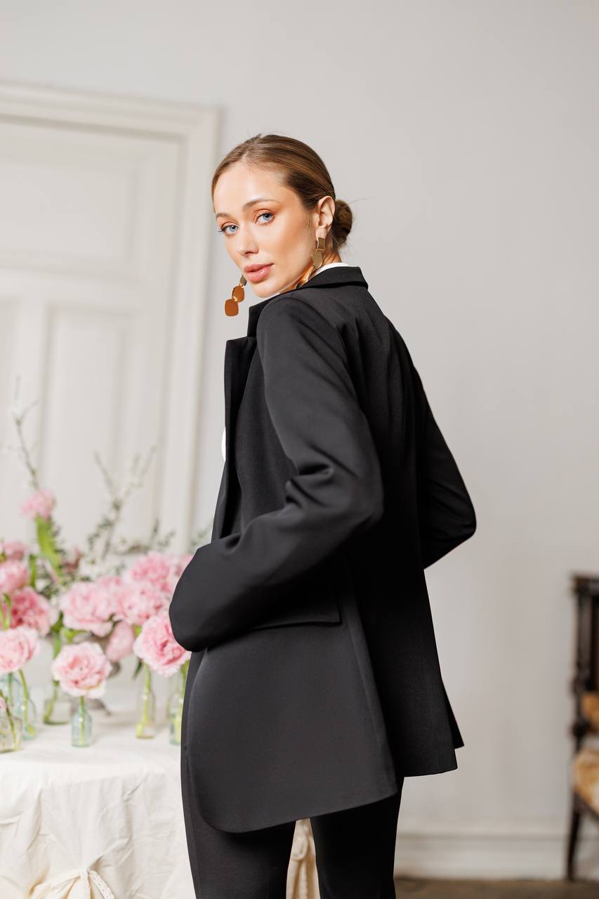 Woman wearing a black blazer in an indoor setting with flowers and furniture.