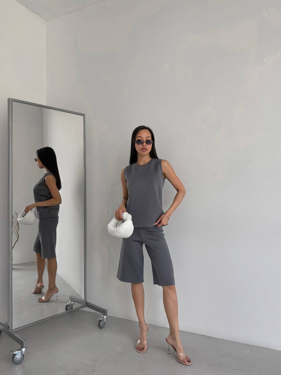 Woman in a gray outfit standing next to a mirror in a minimalistic room.