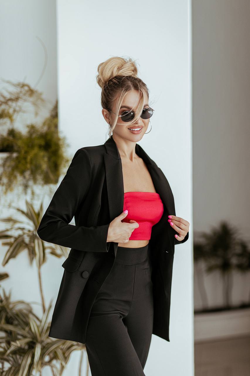 Woman wearing a black coat and red top with sunglasses, standing against a white wall.
