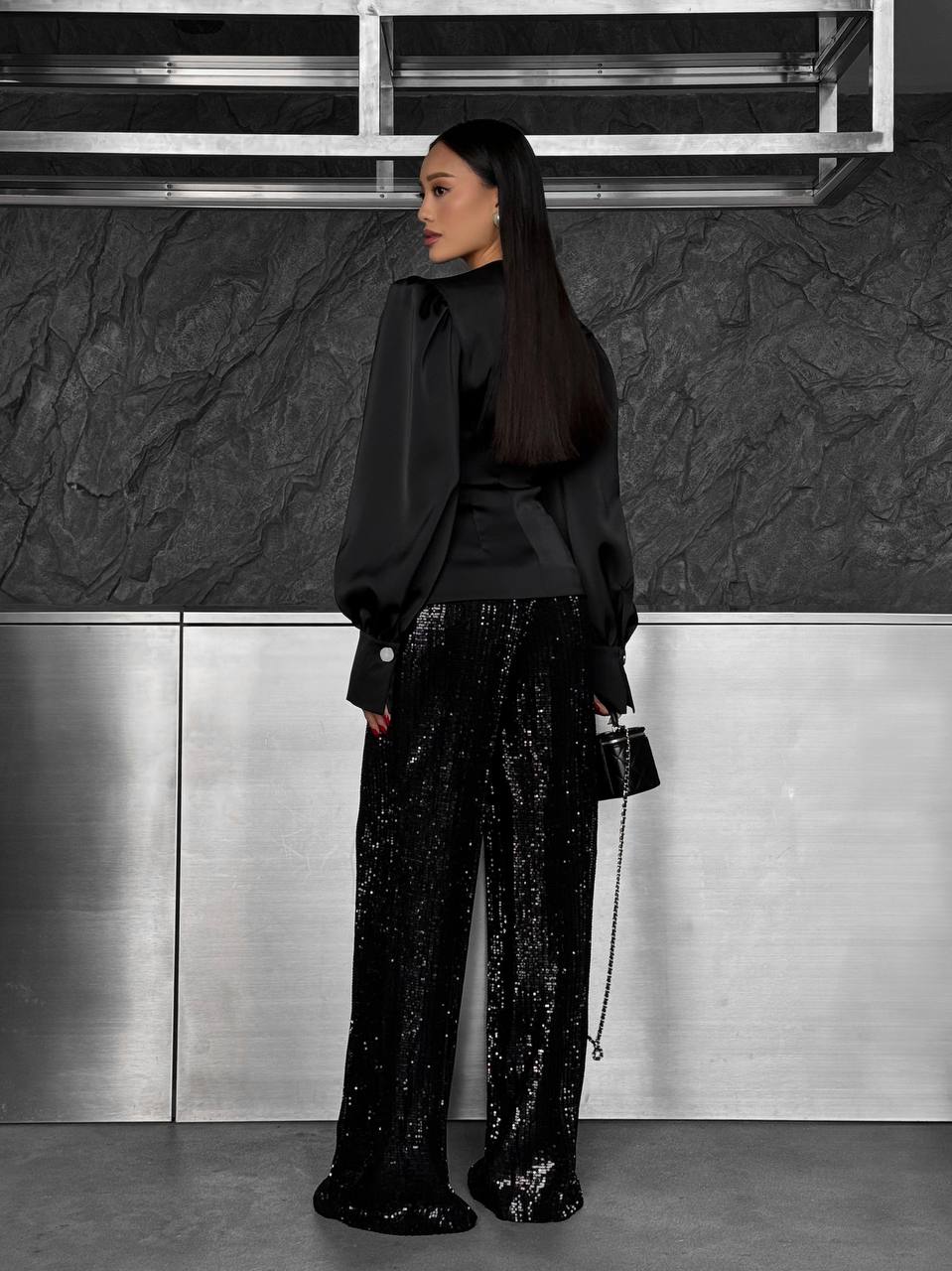 Person wearing a black outfit with sequin pants against a textured wall.