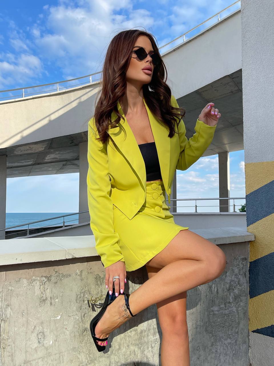 Woman in a bright yellow blazer and black top posing outdoors with ocean view.