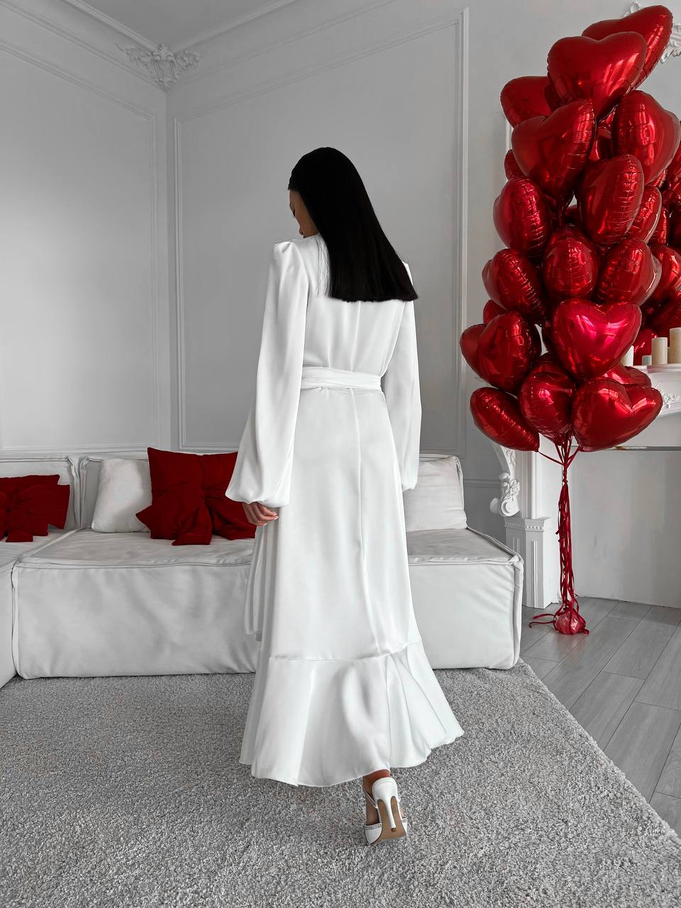 Woman in a white dress standing in a room with red heart-shaped balloons.