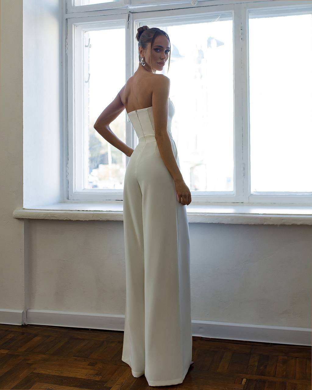 Woman wearing a strapless white jumpsuit standing in front of a window.