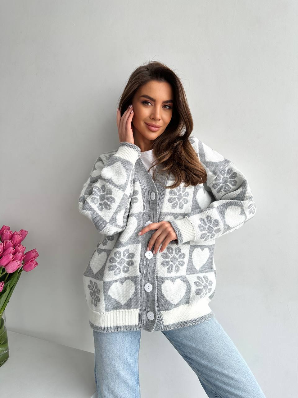 Woman wearing a patterned cardigan with a vase of pink tulips on a light gray background