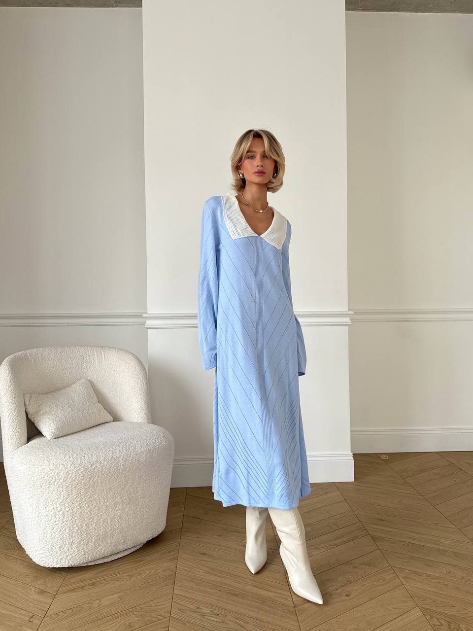Woman in a light blue dress with white collar and cuffs standing in a room with a white armchair.