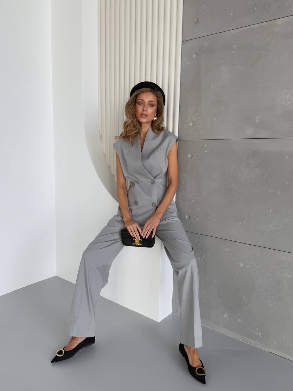 Woman in a gray outfit sitting on a white stool against a concrete wall.