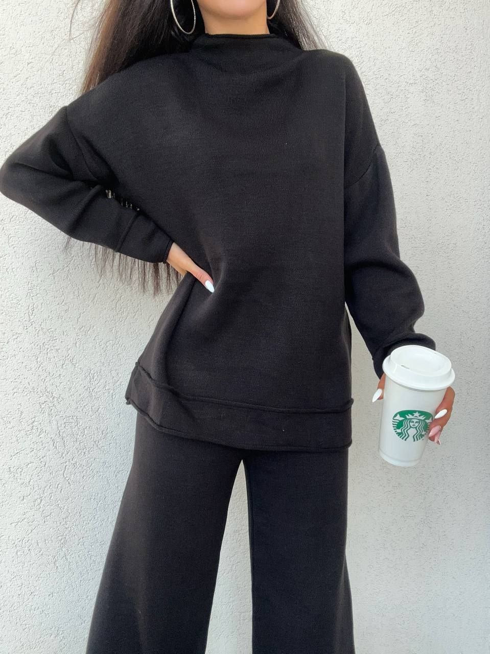 Person wearing a black outfit holding a Starbucks cup against a light background