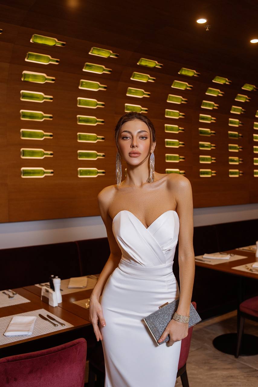 Woman in a white dress standing in a restaurant with a decorative wall of green bottles.