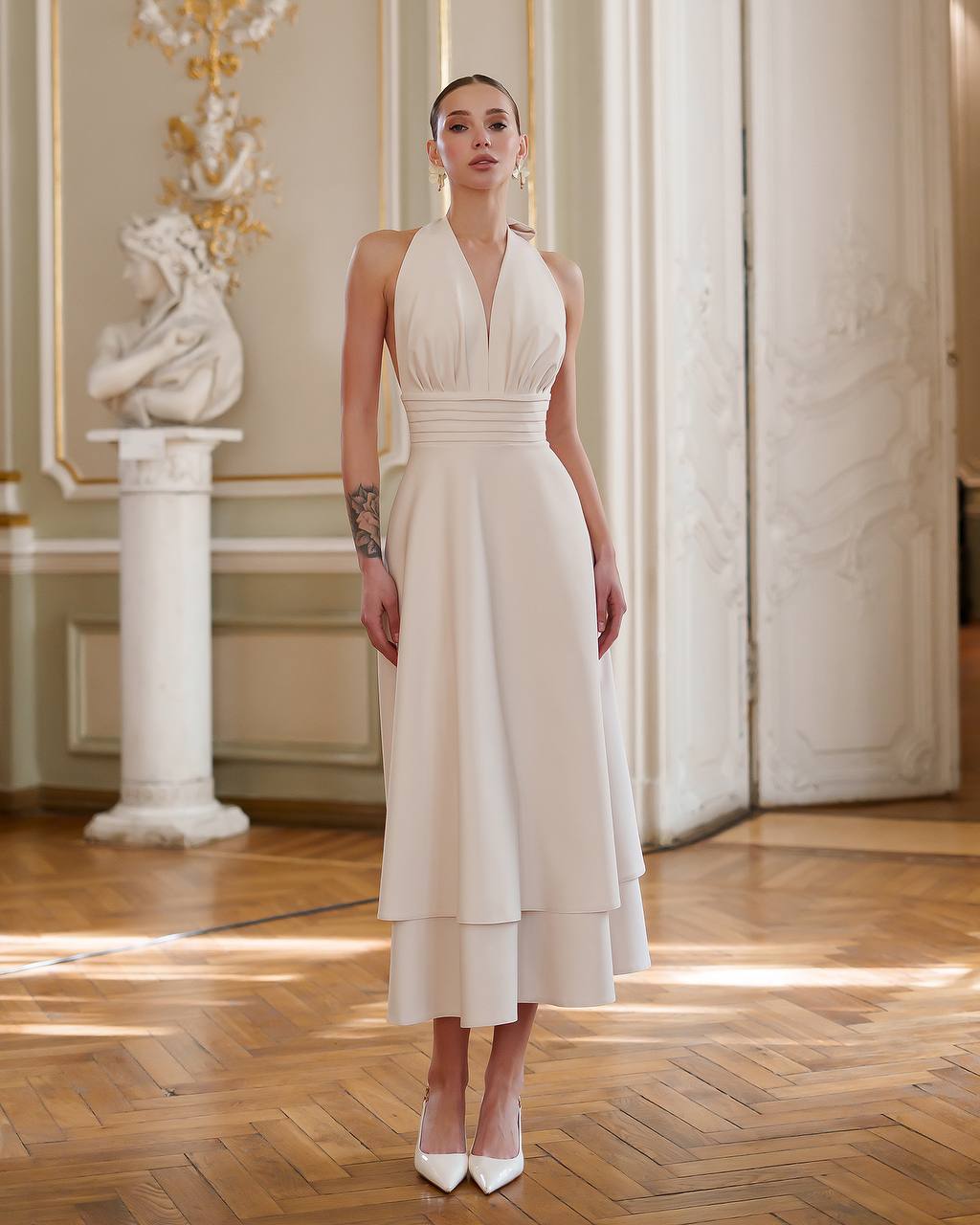 Woman in a white dress standing in an elegant room with marble columns and wooden floor.