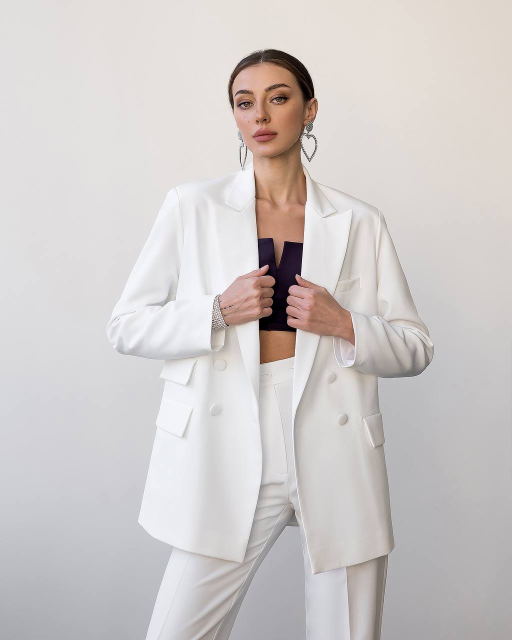 Woman wearing a white blazer over a black top and white pants on a plain background