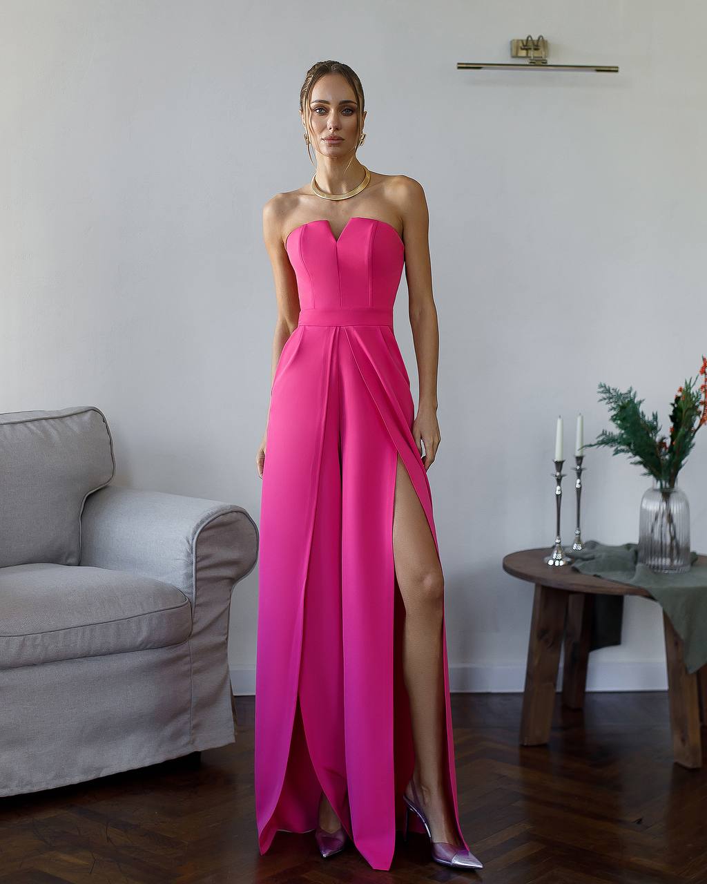 Woman wearing a pink strapless gown with a high slit in a room with a gray sofa and a small table with candles and flowers.