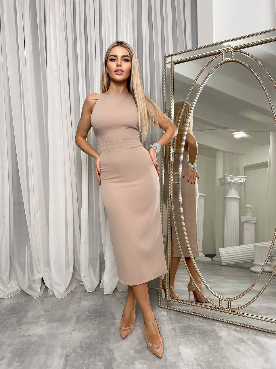 Woman in a beige dress standing in front of a large mirror in a room with white curtains.