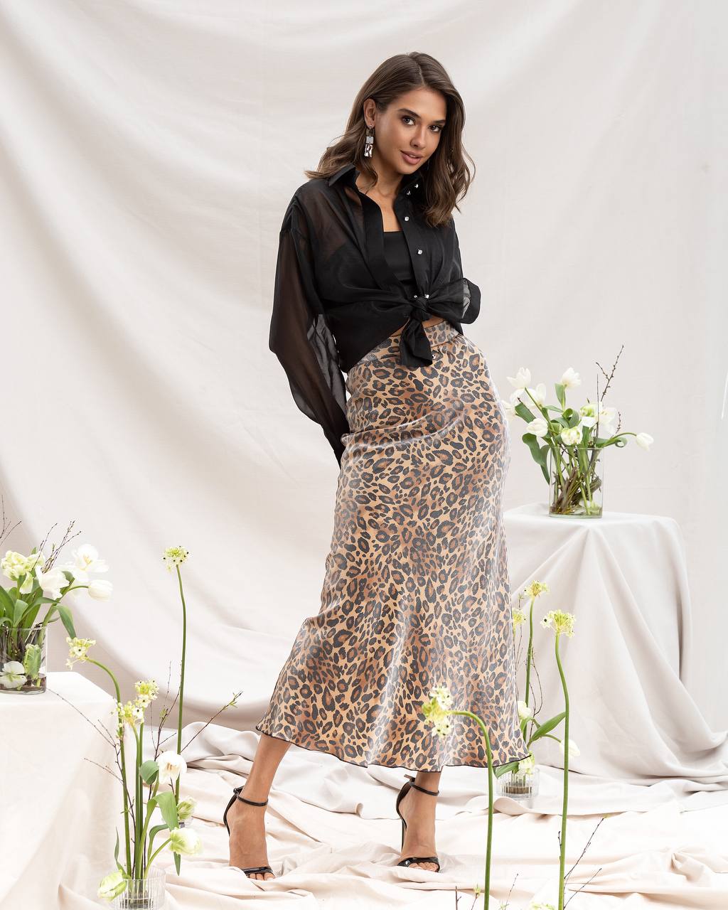 Woman wearing a black blouse and leopard print skirt in a minimalistic setting with flowers.