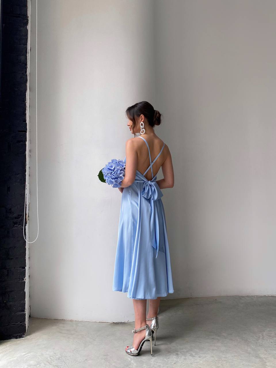Woman in a light blue dress holding flowers against a plain wall.