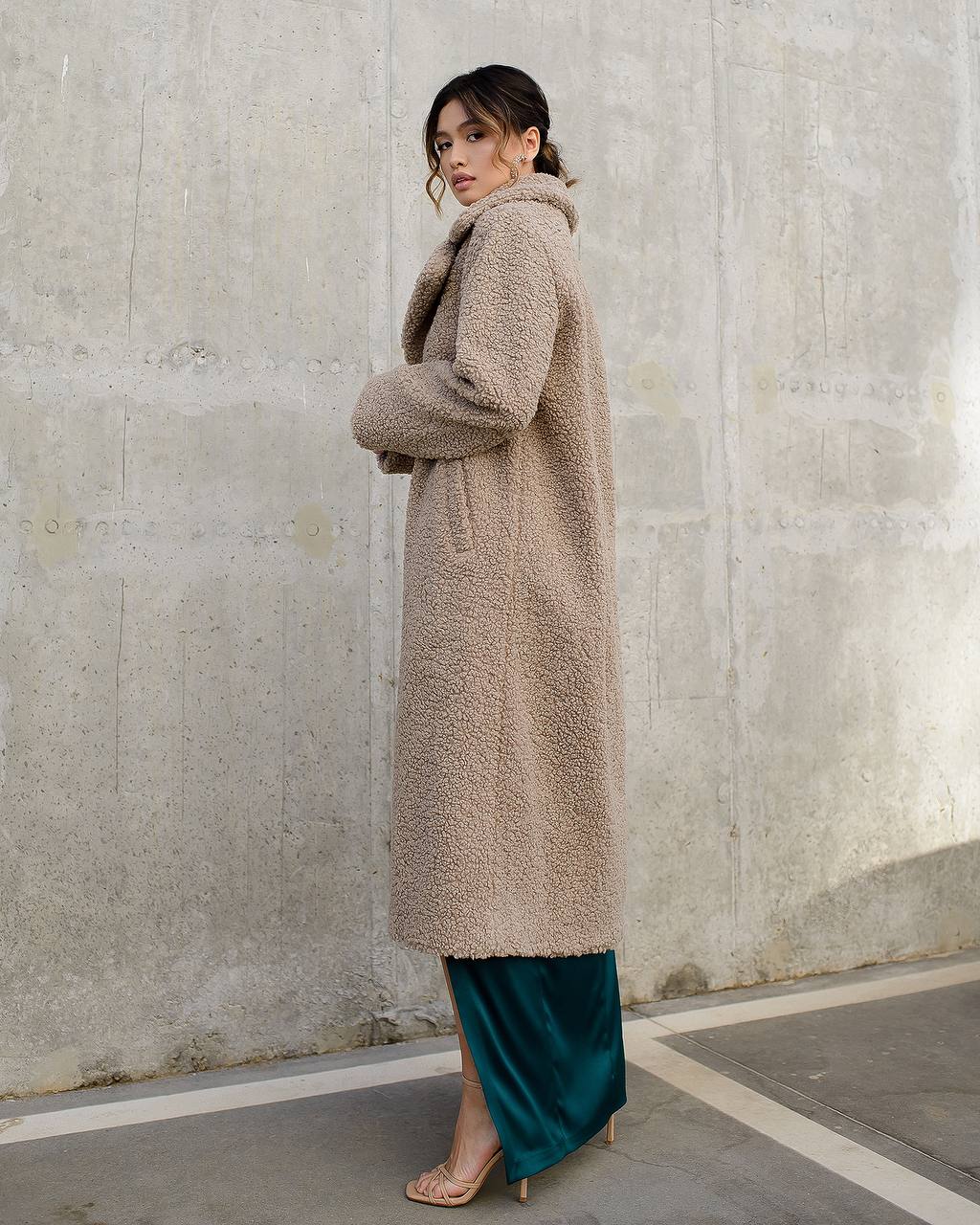 Woman wearing a long beige coat against a concrete wall.