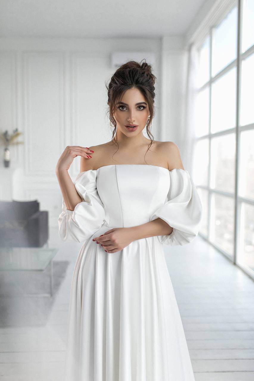 Woman in a white dress standing in a bright room with large windows.