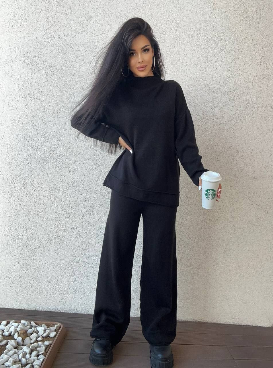 Person wearing a black outfit holding a Starbucks cup against a plain wall.