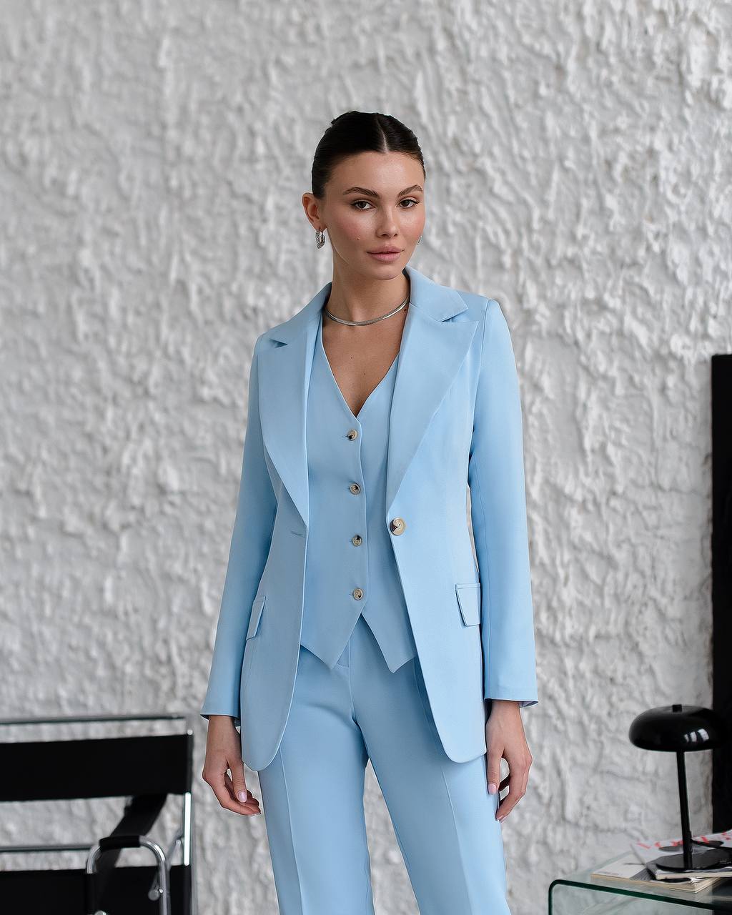 Woman wearing a light blue suit standing against a textured white wall.