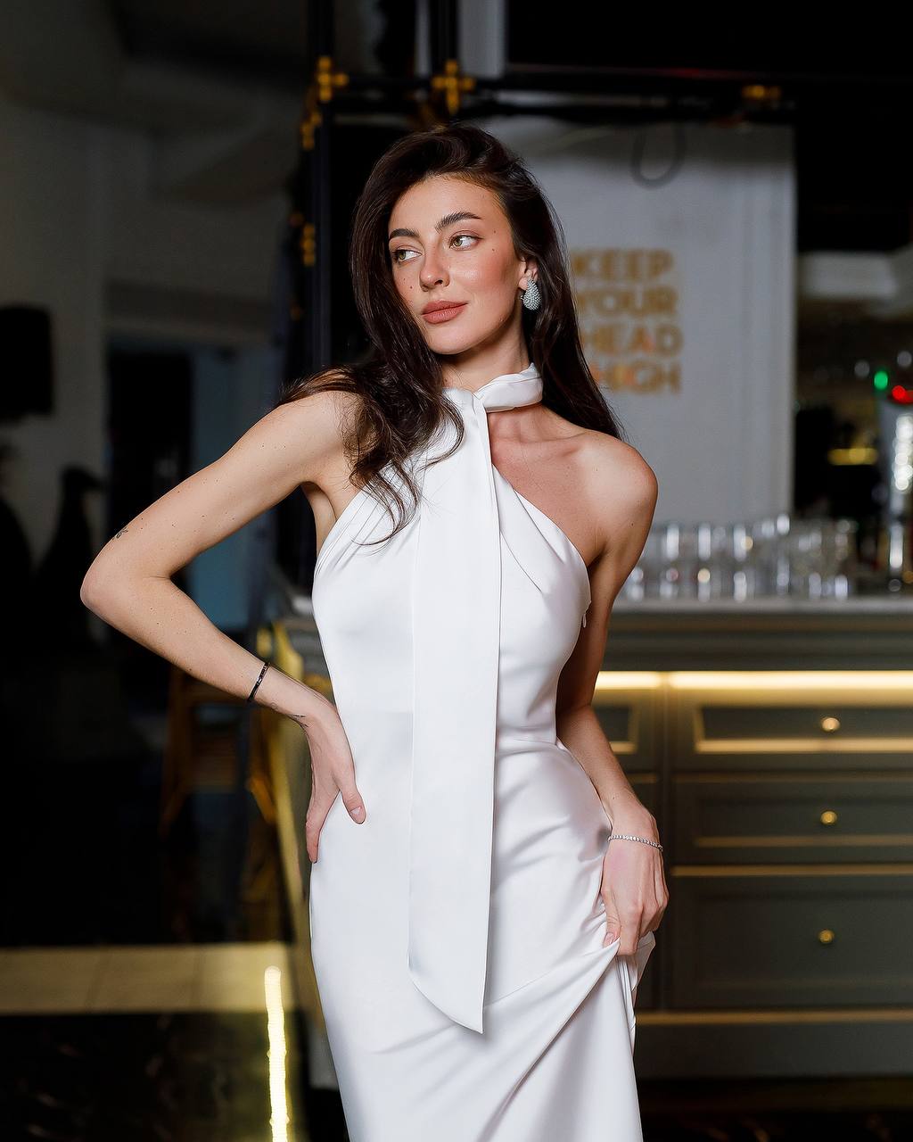 Woman in a white dress posing in an indoor setting with a bar in the background.