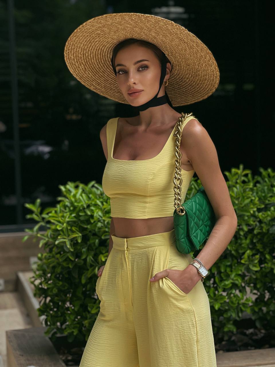Woman in yellow outfit with a wide-brimmed hat and green handbag outdoors.