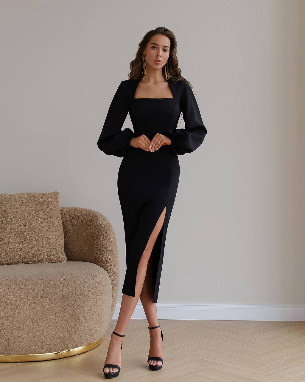 Woman wearing a black dress with a slit standing in a room with a beige armchair.
