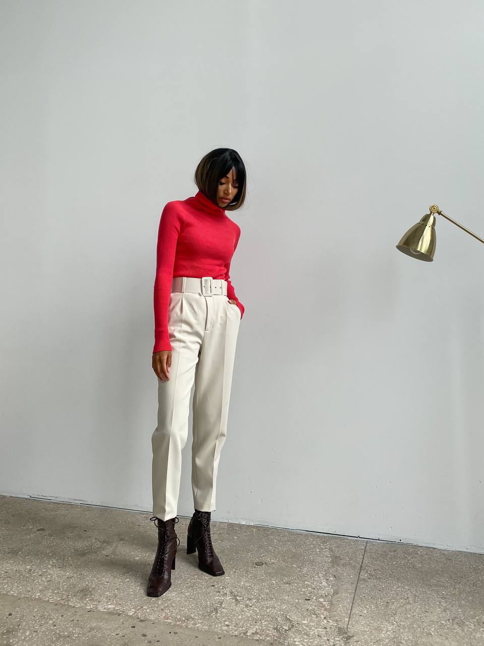 Person wearing a red turtleneck and white pants standing against a plain wall.