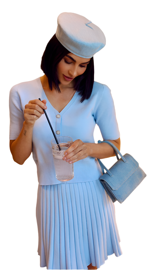 Woman wearing a light blue outfit with a hat and purse, holding a drink.