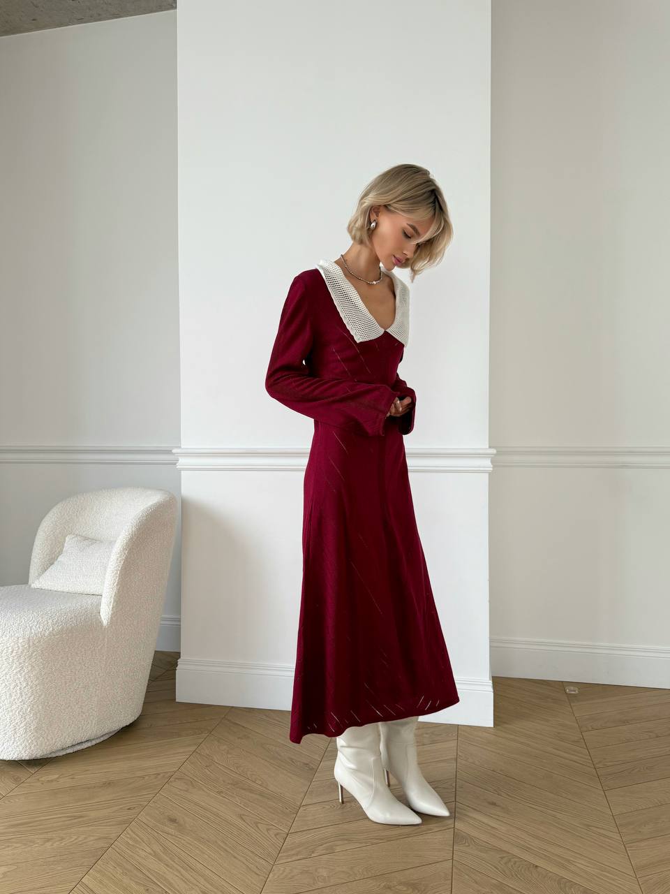 Woman in a burgundy dress with white trim standing in a room with a white wall and wooden floor.