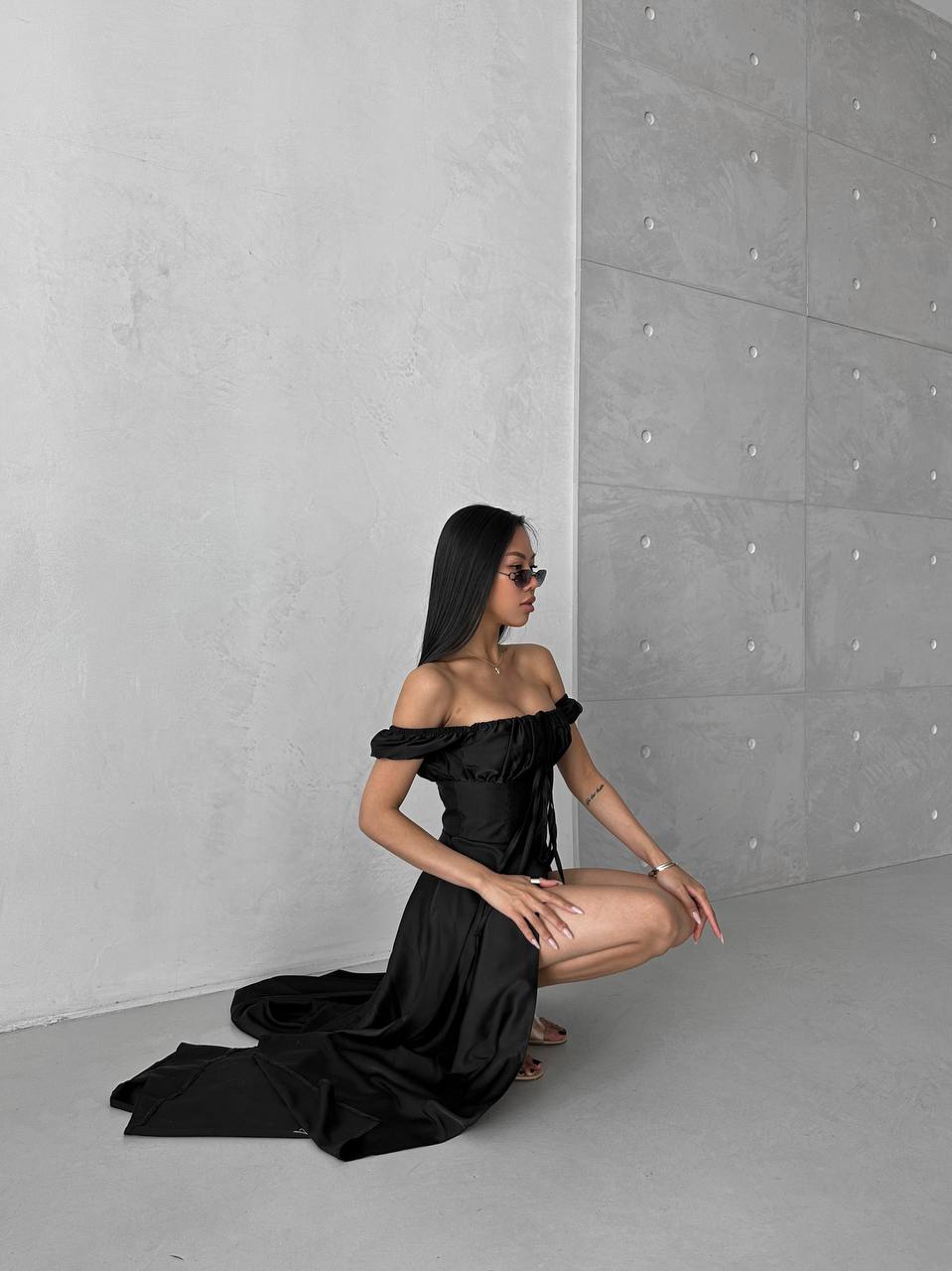 Woman in a black off-shoulder dress posing against a gray concrete wall.
