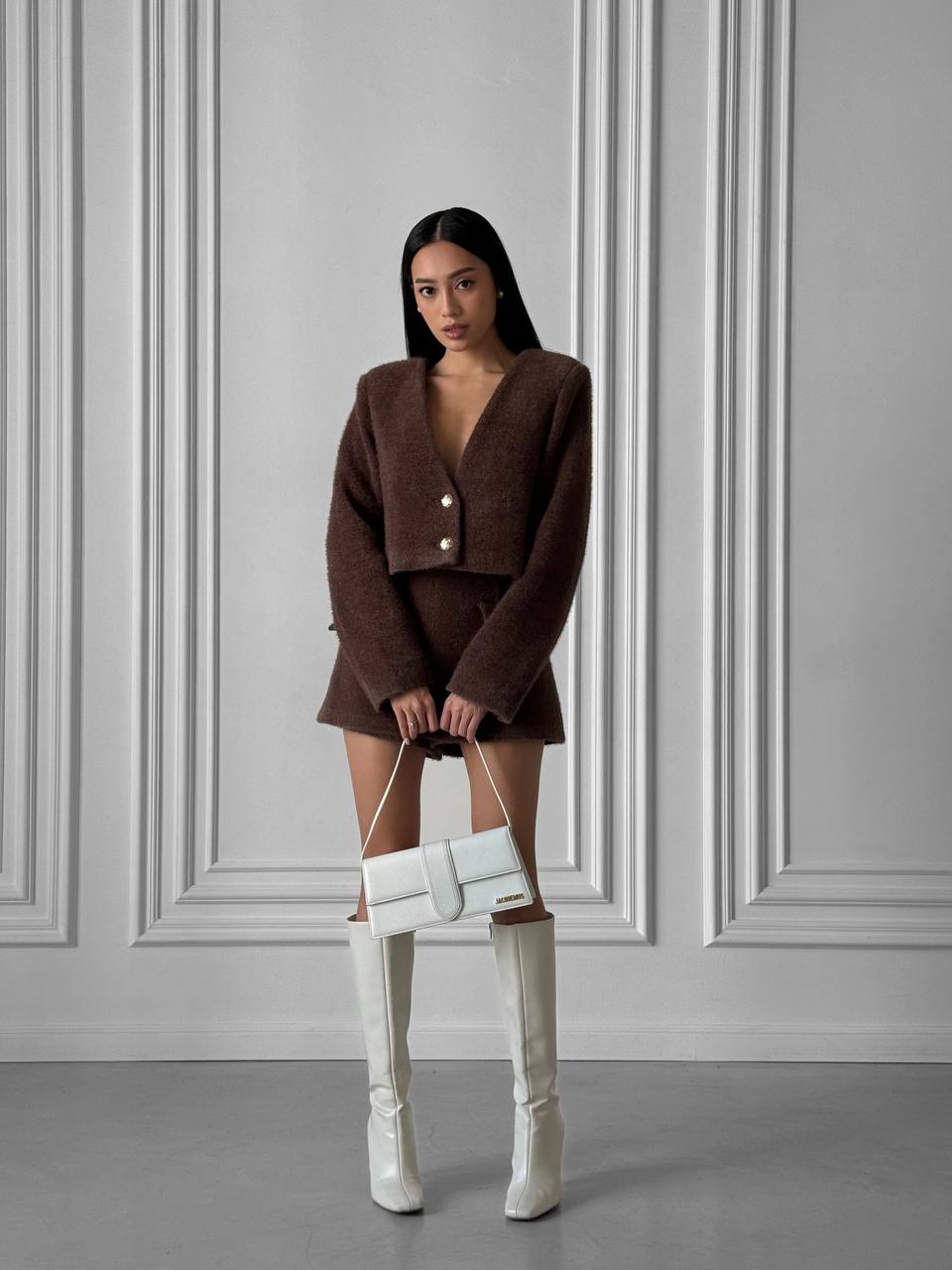 Woman in a brown outfit with white knee-high boots holding a white handbag against a gray wall.