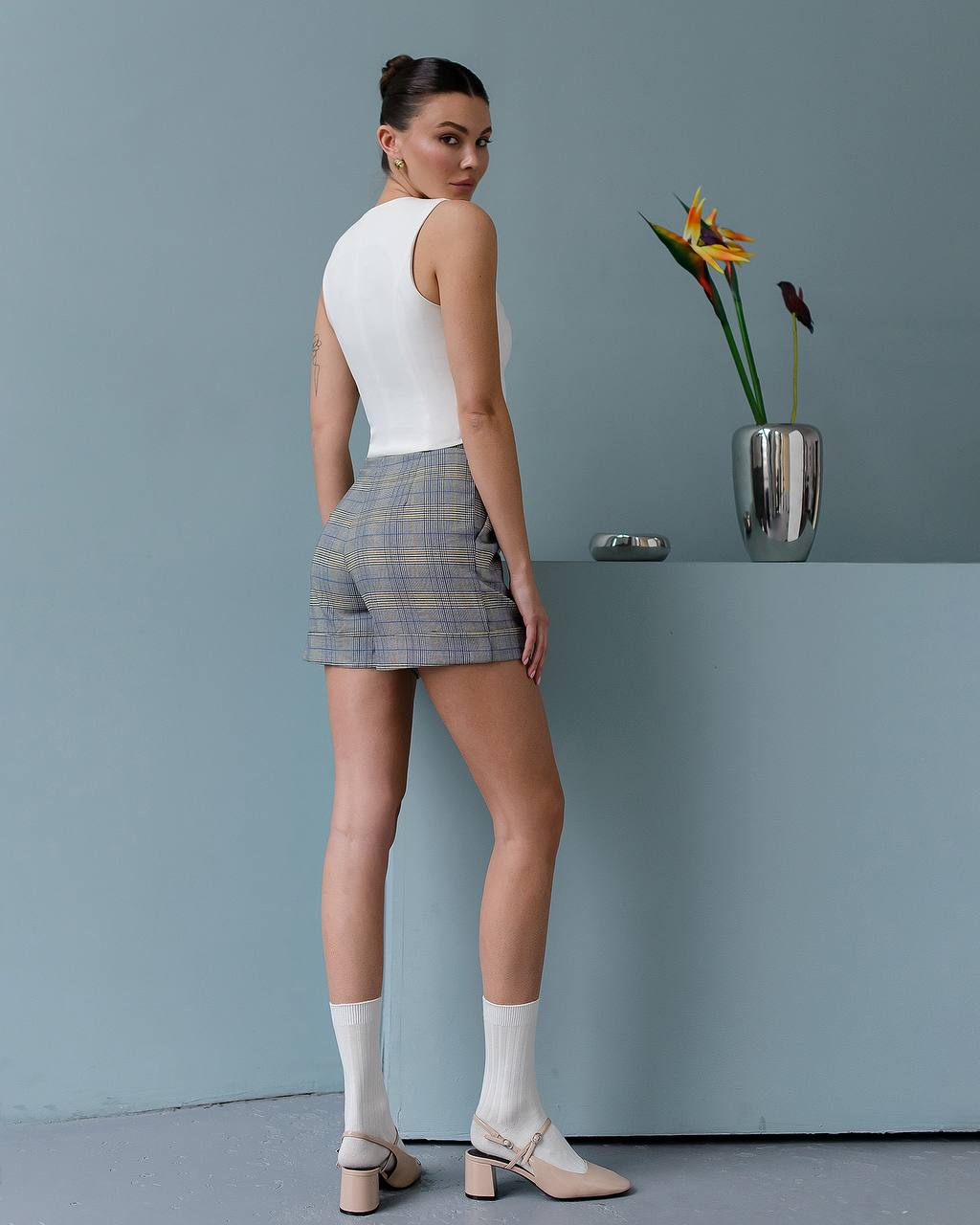 Woman in a white sleeveless top and plaid shorts standing against a plain wall with a vase of flowers.