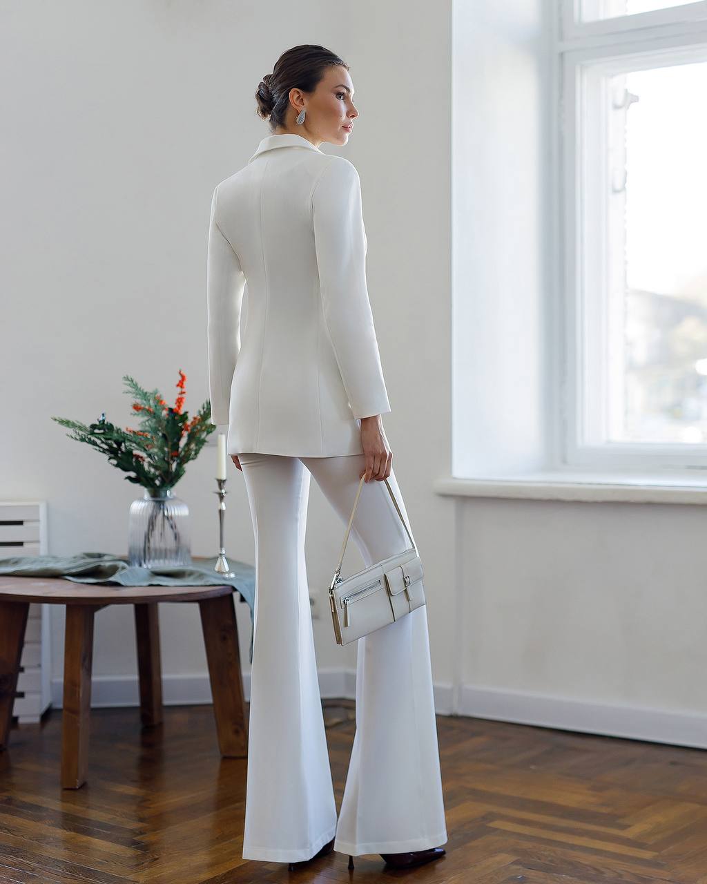 Woman in a white outfit standing in a room with a table and window.