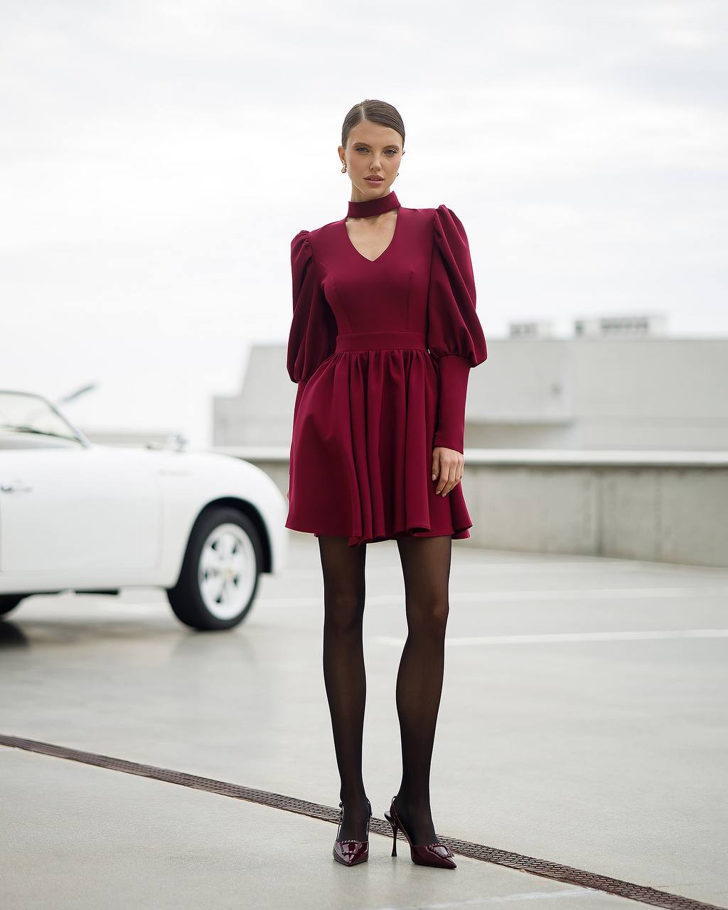 Woman in a burgundy dress standing in a modern indoor setting with a blurred car in the background.