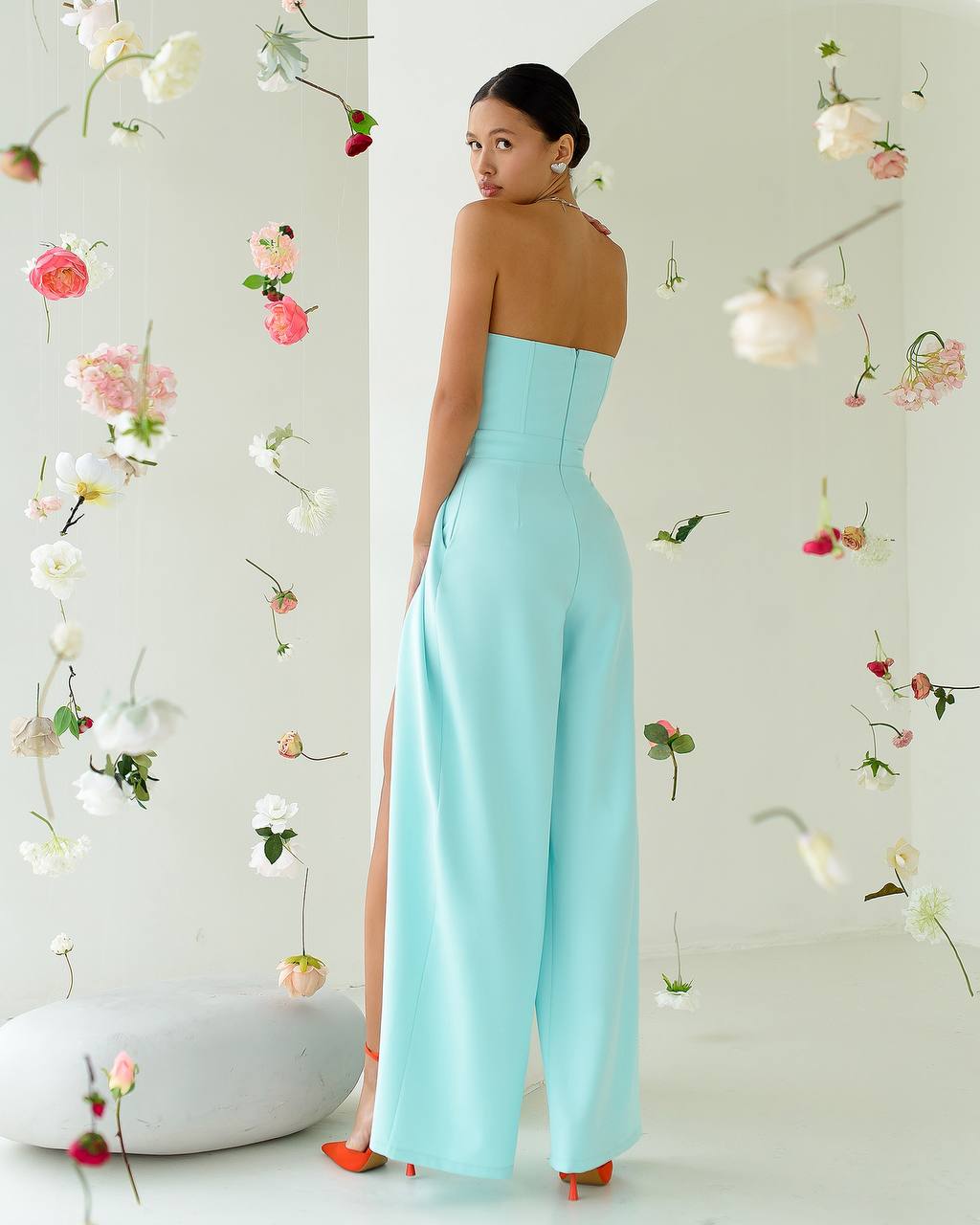 Woman in a light blue strapless jumpsuit with floral decorations on a white wall.