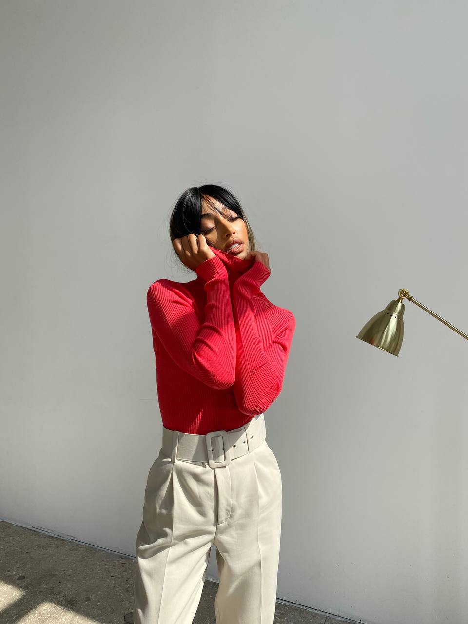 Person wearing a red sweater and beige pants against a plain wall with a gold lamp.