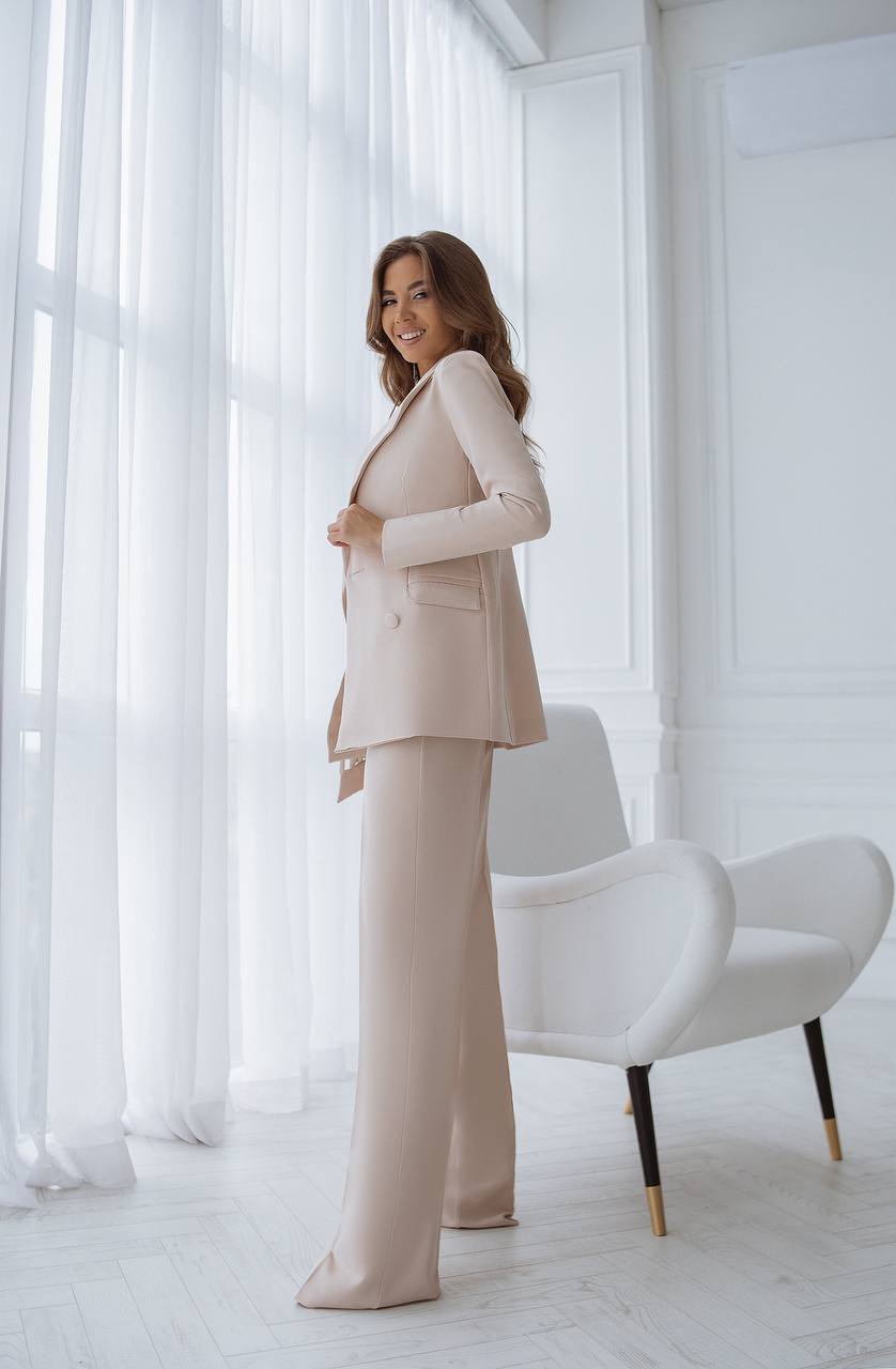 Woman in a beige suit standing in a bright room with white furniture.