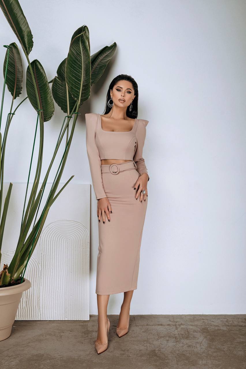 Woman in a beige outfit standing next to a potted plant against a white wall.